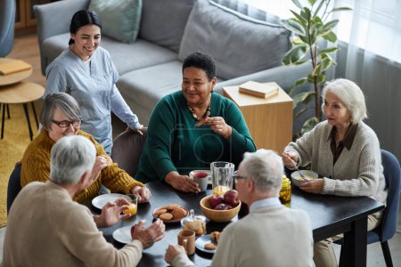 Téléchargez les photos : Vue en angle élevé à un groupe de personnes âgées assises à la table ensemble et souriant joyeusement dans une maison de retraite - en image libre de droit