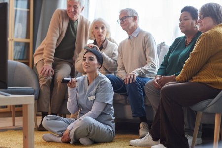 Téléchargez les photos : Groupe diversifié de personnes âgées regardant la télévision dans une maison de retraite avec une jeune infirmière tenant à distance - en image libre de droit