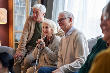 Téléchargez les photos : Vue de côté à un groupe de personnes âgées souriantes regardant la télévision dans une maison de retraite ensemble assis sur un canapé - en image libre de droit