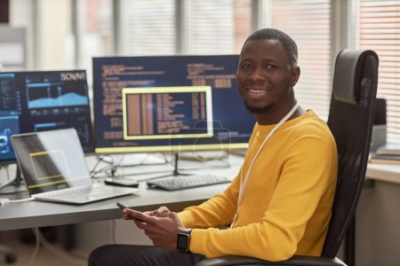 Téléchargez les photos : Portrait d'un homme noir souriant regardant la caméra tout en étant assis sur le lieu de travail dans une société de développement de logiciels contre des écrans d'ordinateur avec du code - en image libre de droit