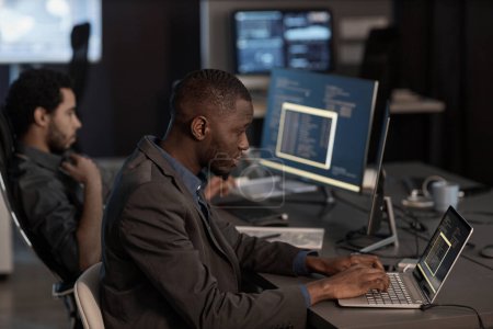 Photo for Side view portrait of black man writing code and typing at laptop keyboard while working in data security office - Royalty Free Image