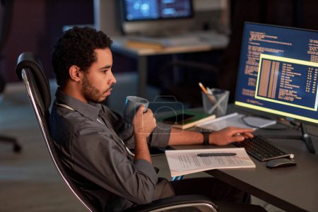 Téléchargez les photos : Portrait latéral du développeur de logiciels masculins écrivant le code et buvant du café tout en travaillant la nuit dans le bureau de sécurité des données - en image libre de droit