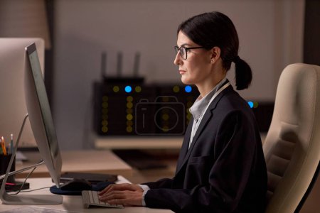 Foto de Retrato de vista lateral de una mujer de negocios seria que usa computadora mientras trabaja por la noche en la oficina de seguridad de datos - Imagen libre de derechos