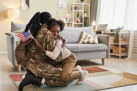 Foto de Niña con bandera americana abrazando a su madre en camuflaje regresando a casa de la guerra - Imagen libre de derechos