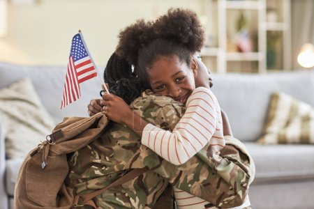 Photo for Portrait of African American little daughter with american flag embracing her mom in military uniform and smiling at camera - Royalty Free Image