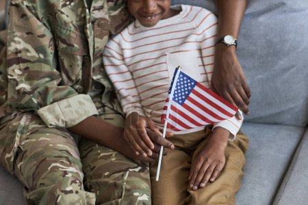Foto de Primer plano de la niña sentada con bandera americana en el sofá y apoyando a su madre en uniforme militar - Imagen libre de derechos