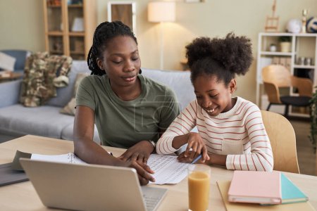 Téléchargez les photos : Mère afro-américaine aider sa fille avec des devoirs alors qu'ils sont assis à table avec ordinateur portable dans le salon - en image libre de droit