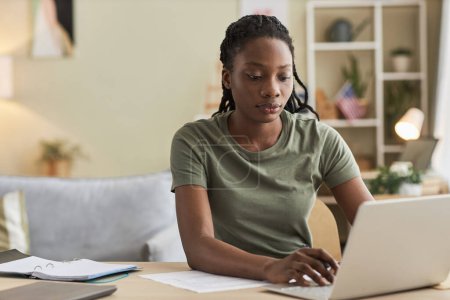 Téléchargez les photos : Afro-américaine jeune femme travaillant en ligne sur ordinateur portable assis à la table dans le salon - en image libre de droit