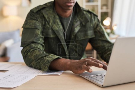 Téléchargez les photos : Gros plan du jeune militaire assis à table à la maison et tapant sur un ordinateur portable, il fait du travail en ligne - en image libre de droit
