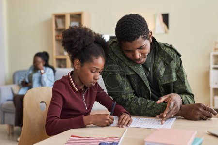Téléchargez les photos : Afro-américaine petite fille faire des devoirs avec son père alors qu'ils assis à la table à la maison - en image libre de droit
