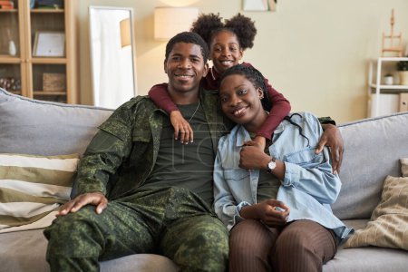 Téléchargez les photos : Portrait de famille afro-américaine de trois personnes souriantes devant une caméra assise sur un canapé avec leur père militaire - en image libre de droit