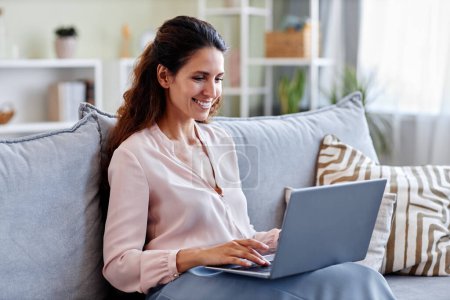 Photo for Portrait of smiling young woman using laptop while sitting on sofa at home and using internet or working online - Royalty Free Image
