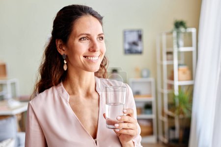 Foto de Retrato de una hermosa mujer adulta bebiendo agua por la mañana mientras comienza el día en casa y sonriendo de pie junto a la ventana - Imagen libre de derechos