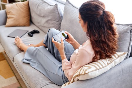 Photo for High angle portrait of young woman playing mobile game via smartphone while relaxing at home - Royalty Free Image