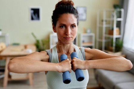 Photo for Front view portrait of young woman holding dumbbells and looking at camera in strength training at home - Royalty Free Image