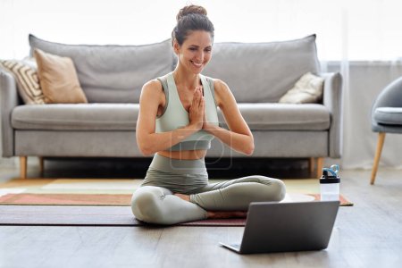 Foto de Retrato de una joven sonriente usando un portátil en el entrenamiento de yoga en casa y meditando con las manos apretadas - Imagen libre de derechos