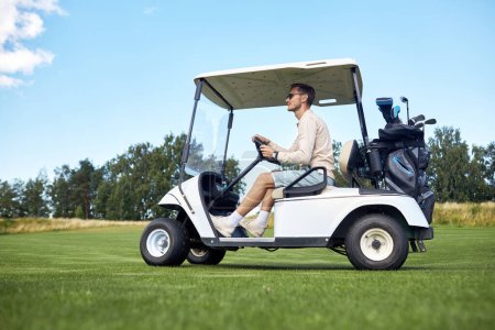 Téléchargez les photos : Vue latérale pleine longueur de l'homme conduisant le chariot de golf à travers le champ vert dans le club de sport, espace de copie - en image libre de droit