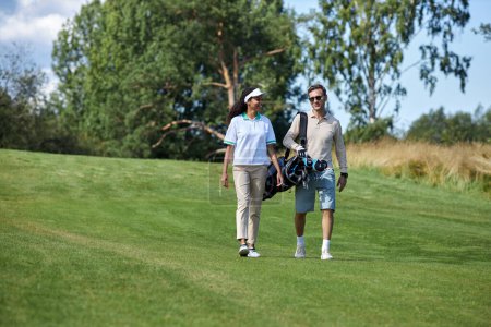 Téléchargez les photos : Portrait complet d'un élégant couple sportif portant un sac de golf marchant sur le champ vert vers la caméra et bavardant, espace de copie - en image libre de droit