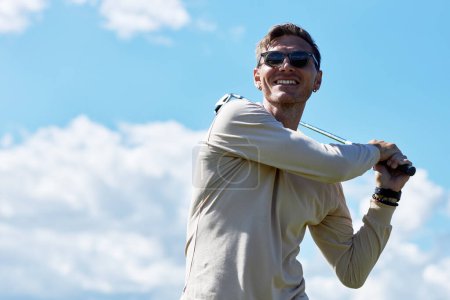 Photo for Waist up portrait of smiling man playing golf and swinging golf club against blue sky outdoors, copy space - Royalty Free Image