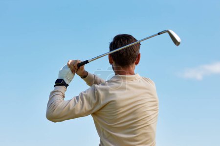 Téléchargez les photos : Vue arrière de l'homme méconnaissable balançant club de golf contre ciel bleu clair - en image libre de droit
