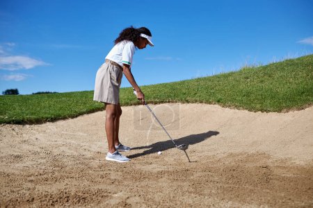 Foto de Retrato de larga duración de una joven negra jugando al golf al aire libre parada en arenero y apuntando al tiro - Imagen libre de derechos