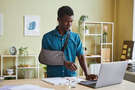 Photo for Portrait of black young man with arm sling working at standing desk and using laptop in office - Royalty Free Image