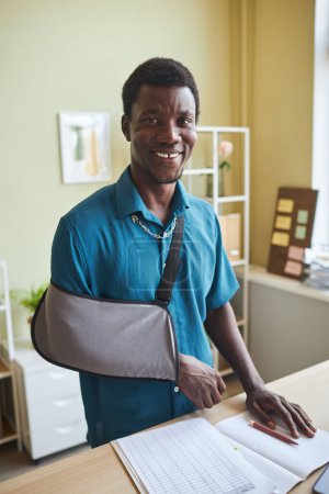 Photo for Vertical portrait of black young man with arm sling working at standing desk and smiling at camera - Royalty Free Image