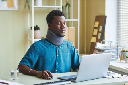 Téléchargez les photos : Portrait de jeune homme noir avec attelle de cou travaillant au bureau et utilisant un ordinateur portable - en image libre de droit
