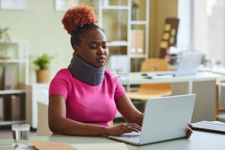 Téléchargez les photos : Portrait de jeune femme noire avec attelle de cou travaillant au bureau et utilisant un ordinateur portable - en image libre de droit