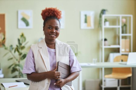 Photo for Waist up portrait of black woman with natural curly hair smiling at camera while standing in pastel colored office, copy space - Royalty Free Image