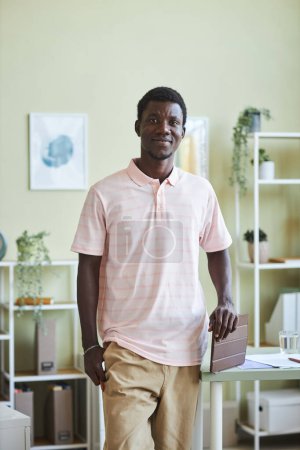 Photo for Vertical portrait of young black man smiling at camera standing by desk in pastel colored office - Royalty Free Image