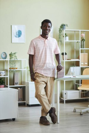 Photo for Vertical full length portrait of young black man smiling at camera standing by desk in cozy office - Royalty Free Image