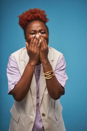 Photo for Candid portrait of emotional black woman posing against vibrant blue background in studio and covering mouth or screaming - Royalty Free Image
