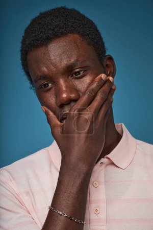 Foto de Retrato vertical de primer plano del hombre negro angustiado cubriendo la boca contra el fondo azul - Imagen libre de derechos