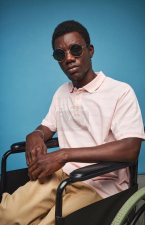 Téléchargez les photos : Portrait vertical de l'homme noir à la mode avec handicap portant des lunettes de soleil sur fond bleu vif - en image libre de droit
