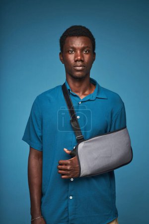 Téléchargez les photos : Portrait vertical du jeune homme noir portant une fronde de bras sur fond bleu vif et regardant la caméra avec expression du visage vide - en image libre de droit