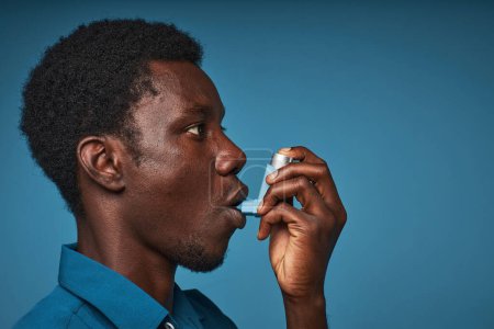 Téléchargez les photos : Vue latérale minime portrait de jeune homme noir utilisant un inhalateur d'asthme sur fond bleu, espace de copie - en image libre de droit