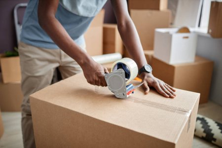 Photo for Close-up of African American man using adhesive tape to pack things into cardboard boxes for relocation - Royalty Free Image