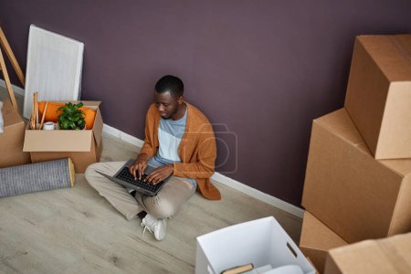 Téléchargez les photos : Afro-Américain assis sur le sol parmi les boîtes et tapant sur ordinateur portable pendant la relocalisation - en image libre de droit