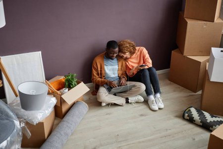 Photo for High angle view of multiethnic young couple using laptop together while sitting on the floor among boxes - Royalty Free Image