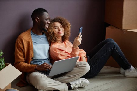 Photo for Young woman showing photos on her smartphone to young man while he using laptop, they sitting on the floor in the room - Royalty Free Image