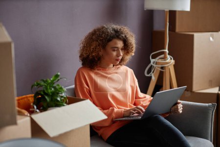 Téléchargez les photos : Jeune femme aux cheveux bouclés assise sur un canapé et tapant sur un ordinateur portable, elle travaille en ligne à la maison - en image libre de droit