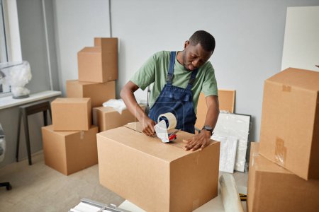 Téléchargez les photos : Travailleur afro-américain dans des choses uniformes d'emballage dans des boîtes en carton avec du ruban adhésif pour la relocalisation - en image libre de droit