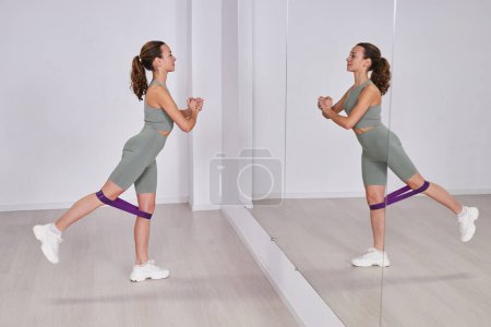 Téléchargez les photos : Jeune femme en bonne santé debout devant le miroir et l'entraînement avec élastique pendant l'entraînement dans la salle de gym - en image libre de droit