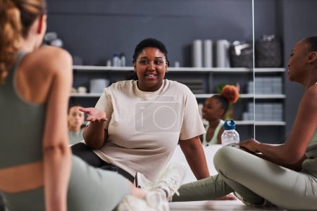 Foto de Grupo de mujeres jóvenes sentadas en el suelo y hablando entre sí durante el entrenamiento en el gimnasio - Imagen libre de derechos