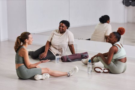 Foto de Mujeres jóvenes sentadas en el suelo y hablando con el instructor sobre el entrenamiento en el gimnasio - Imagen libre de derechos