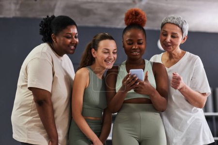 Téléchargez les photos : Groupe de femmes multiethniques regardant des photos sur smartphone tout en étant debout dans un club de santé après l'entraînement - en image libre de droit