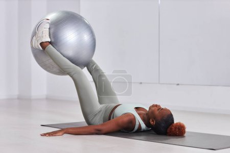 Foto de Mujer joven afroamericana acostada en una colchoneta y ejercitándose con una pelota de fitness - Imagen libre de derechos