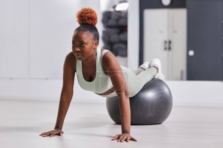 Téléchargez les photos : Afro-Américaine jeune fille en vêtements de sport formation sur la balle de fitness dans la salle de gym - en image libre de droit