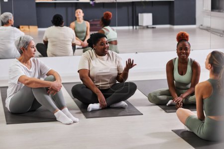 Téléchargez les photos : Femmes assises sur le tapis d'exercice et discutant de la formation avec l'entraîneur dans un club de santé - en image libre de droit